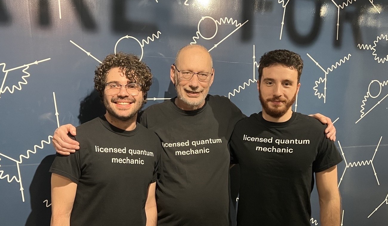 Samuel Patrone, Mark Wise, and Adriano Testa smiling together in front of a Feynman-diagram-covered wall, all three wearing t-shirts reading "licensed quantum mechanic"