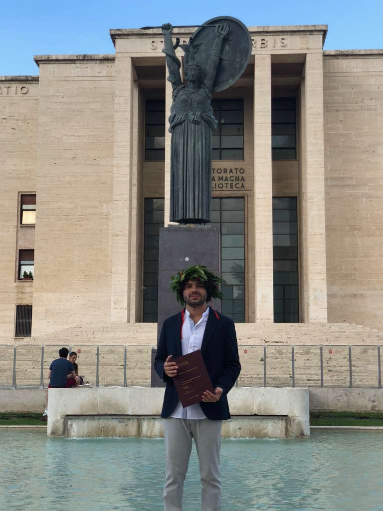 Samuel Patrone with a laurel wreath holding his thesis in front of the Minerva statue at Sapienza University of Rome